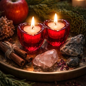 May include: Two heart-shaped red glass candle holders with lit white candles, displayed on a brass tray with cinnamon sticks, a red apple, crystals, and greenery. The scene evokes a sense of warmth and tranquility.