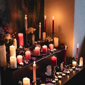 May include: A display of lit candles in shades of red, pink, and white, on a dark wooden surface. Some candles are in holders, with rose petals and crystals. The scene is dimly lit, creating a warm and inviting atmosphere.