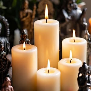 Pode incluir: Cinco velas de pilar de cor marfim com chamas acesas. As velas de diferentes alturas s&atilde;o cercadas por pequenas est&aacute;tuas, cristais e pedras. O fundo est&aacute; desfocado, sugerindo um tema espiritual ou decorativo.