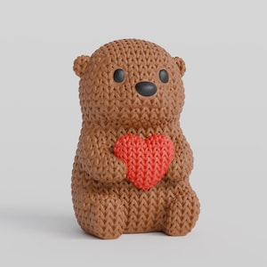 Könnte beinhalten: Eine braune, gestrickte Bärenfigur, die ein rotes Herz hält. Der Bär hat schwarze Augen und eine schwarze Nase. Die Figur besteht aus einem strukturierten Material, das ihr ein handgemachtes Aussehen verleiht. Der Hintergrund ist einfarbig und neutral.