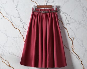 May include: A burgundy midi skirt with a matching belt, hanging on a wooden hanger. The skirt has a pleated design and a cinched waist. The belt is a woven style with a silver-tone buckle. The background is a marble wall.