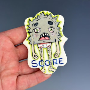 May include: A ceramic tile with a cartoon character with a gray body, green eyes, and a red mouth. The character is wearing white underwear and has hairy legs. The tile is decorated with yellow and green accents and the word "SCORE" in blue letters.