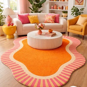 May include: A vibrant orange rug with a unique, wavy shape and a pink and beige striped border. The rug is in a living room with a white sofa, yellow armchair, and a white coffee table. The room has a bright and colourful aesthetic.