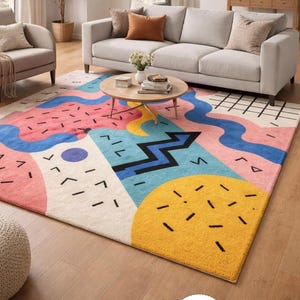 Op de afbeelding: Een kleurrijk vloerkleed met een geometrisch ontwerp. Het vloerkleed heeft een mix van roze, blauwe, gele en witte vormen met zwarte accenten. Het vloerkleed ligt in een woonkamer.