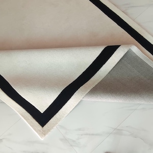 Peut inclure: Un tapis rectangulaire présentant une surface de couleur crème avec une bordure noire marquée. Un coin du tapis est replié pour révéler le dessous, qui affiche un matériau gris clair texturé correspondant à la structure du dos du tapis.