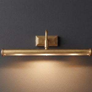 May include: A brass-toned picture light fixture. The rectangular backplate supports a curved arm and a horizontal light bar with rounded end caps. The light is on, casting a warm glow on the wall below.