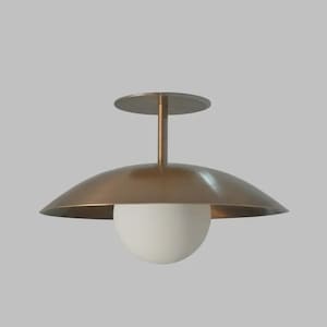 Puede incluir: Una moderna lámpara de techo con una pantalla en forma de platillo de color latón y un globo de cristal blanco esférico. La luz está suspendida de un soporte de techo circular mediante una fina varilla.