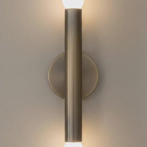 May include: A contemporary wall light with a cylindrical, brushed metal body and a circular backplate. It has a light bulb at the top and bottom, providing a warm light. The fixture is designed for indoor use.