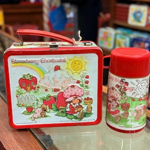 May include: A vintage Strawberry Shortcake lunchbox and matching flask. The red metal lunchbox features a colourful illustration of Strawberry Shortcake and friends. The flask has a red lid and a similar design.