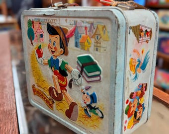 May include: A vintage Pinocchio lunchbox with a red handle. The lunchbox features colourful illustrations of Pinocchio, Jiminy Cricket, and other characters. The lunchbox has a white background with a silver trim.