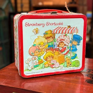 May include: A vintage Strawberry Shortcake lunchbox with a red handle and trim. The front panel displays a colourful illustration of Strawberry Shortcake and friends at a market stall. A nostalgic collectible.