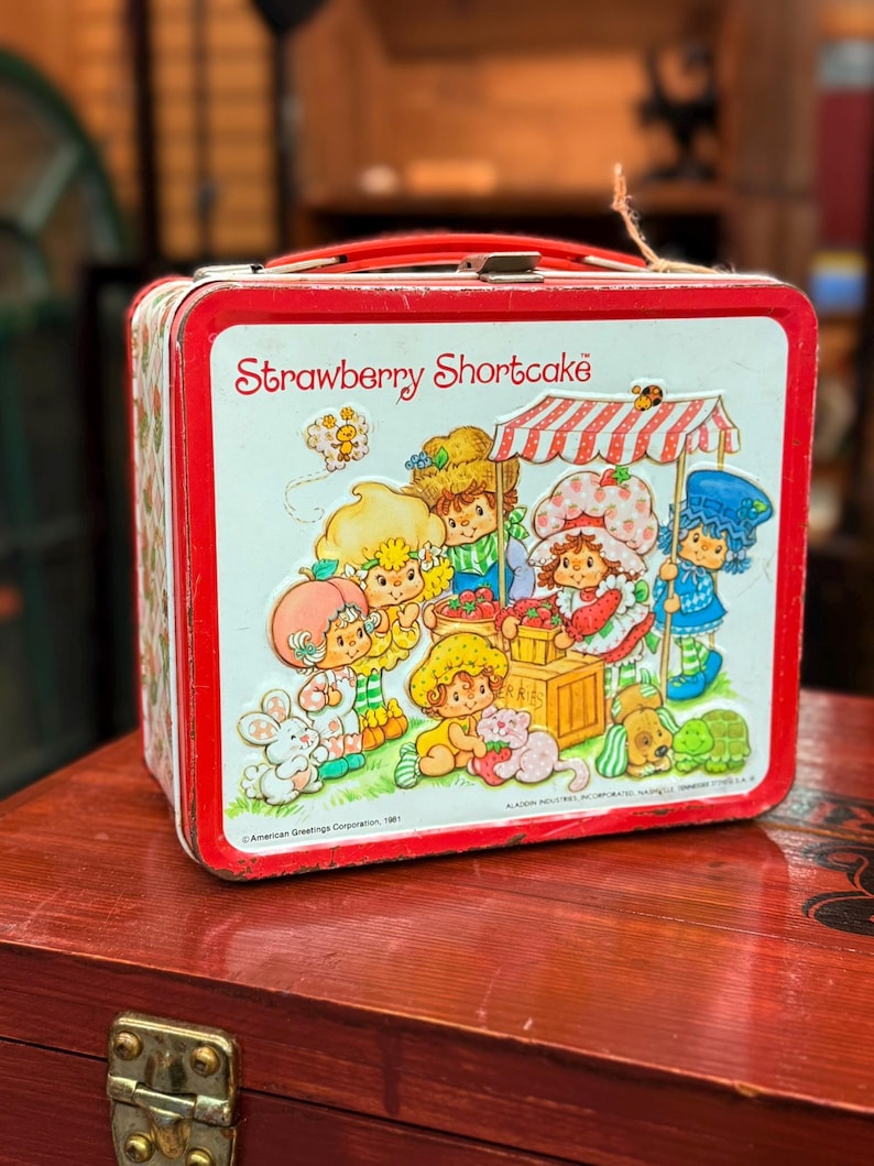 May include: A vintage Strawberry Shortcake lunchbox with a red handle and trim. The front panel displays a colourful illustration of Strawberry Shortcake and friends at a market stall. A nostalgic collectible.