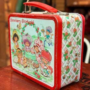 May include: A vintage Strawberry Shortcake lunchbox with a red handle and trim. The lunchbox features illustrations of Strawberry Shortcake characters, with a repeating pattern of strawberries and flowers on the sides.