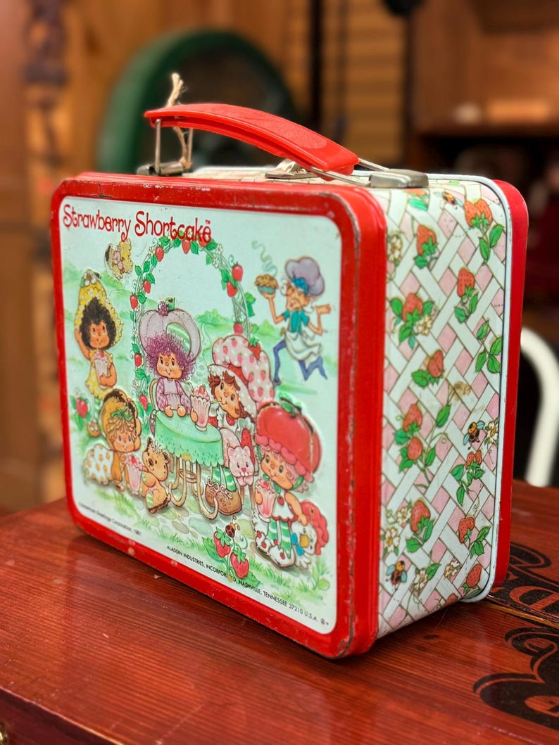 May include: A vintage Strawberry Shortcake lunchbox with a red handle and trim. The lunchbox features illustrations of Strawberry Shortcake characters, with a repeating pattern of strawberries and flowers on the sides.