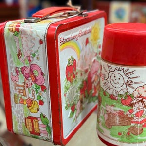 May include: A vintage Strawberry Shortcake lunchbox and flask set. The lunchbox is red with a white panel featuring Strawberry Shortcake characters. The flask is red and white, with a matching design. A nostalgic collectible item.