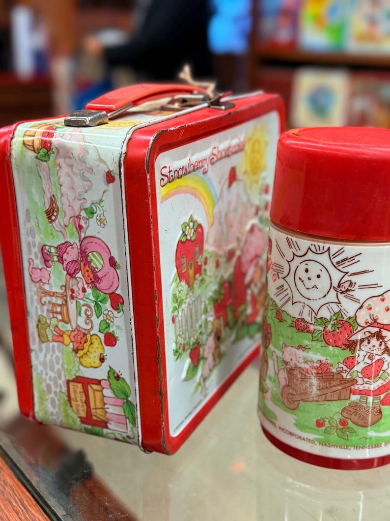 May include: A vintage Strawberry Shortcake lunchbox and flask set. The lunchbox is red with a white panel featuring Strawberry Shortcake characters. The flask is red and white, with a matching design. A nostalgic collectible item.
