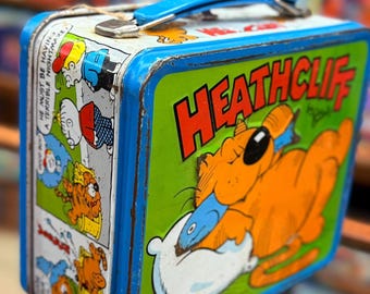 May include: Vintage Heathcliff lunchbox with a blue handle and trim. The lunchbox features a cartoon image of Heathcliff the cat on a green background, biting a pillow. The side panels show comic strip panels.