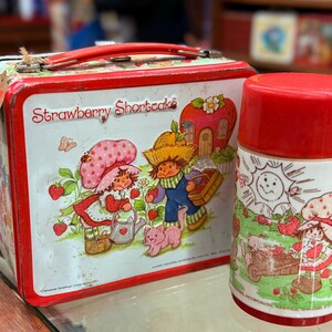 May include: A vintage Strawberry Shortcake lunchbox and flask set. The lunchbox is red and white with a handle and features illustrations of Strawberry Shortcake characters. The flask has a red lid and a matching design. The set is from 1980.