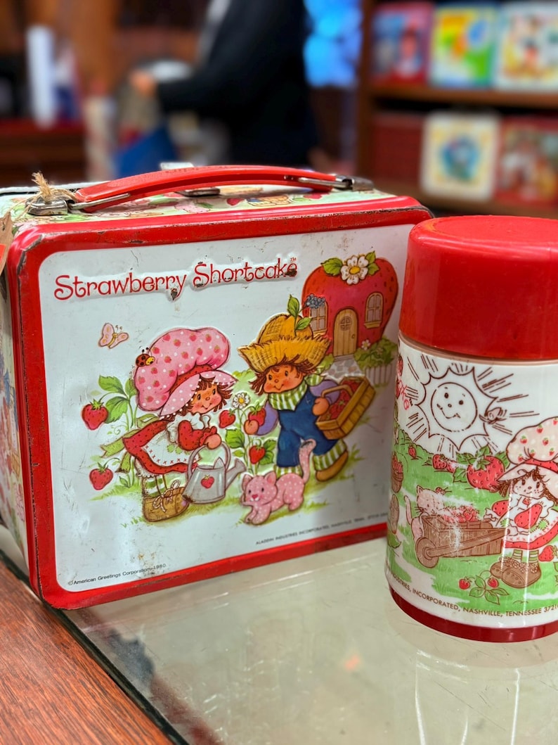 May include: A vintage Strawberry Shortcake lunchbox and flask set. The lunchbox is red and white with a handle and features illustrations of Strawberry Shortcake characters. The flask has a red lid and a matching design. The set is from 1980.