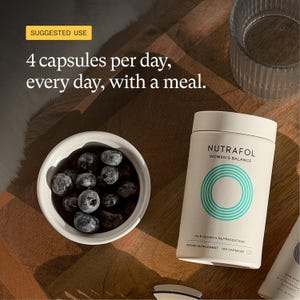 May include: A white cylindrical bottle of Nutrafol Women's Balance hair growth supplement with a teal circular design and text. A small white bowl filled with blueberries sits next to the bottle. The text "4 capsules per day, every day, with a meal" is also visible.