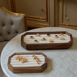 Pode incluir: Duas bandejas de joias de madeira com interior de cor clara, exibindo vários anéis e brincos. Os anéis apresentam diferentes pedras preciosas e designs, incluindo faixas de ouro e pedras coloridas. As bandejas estão sobre uma superfície semelhante ao mármore.