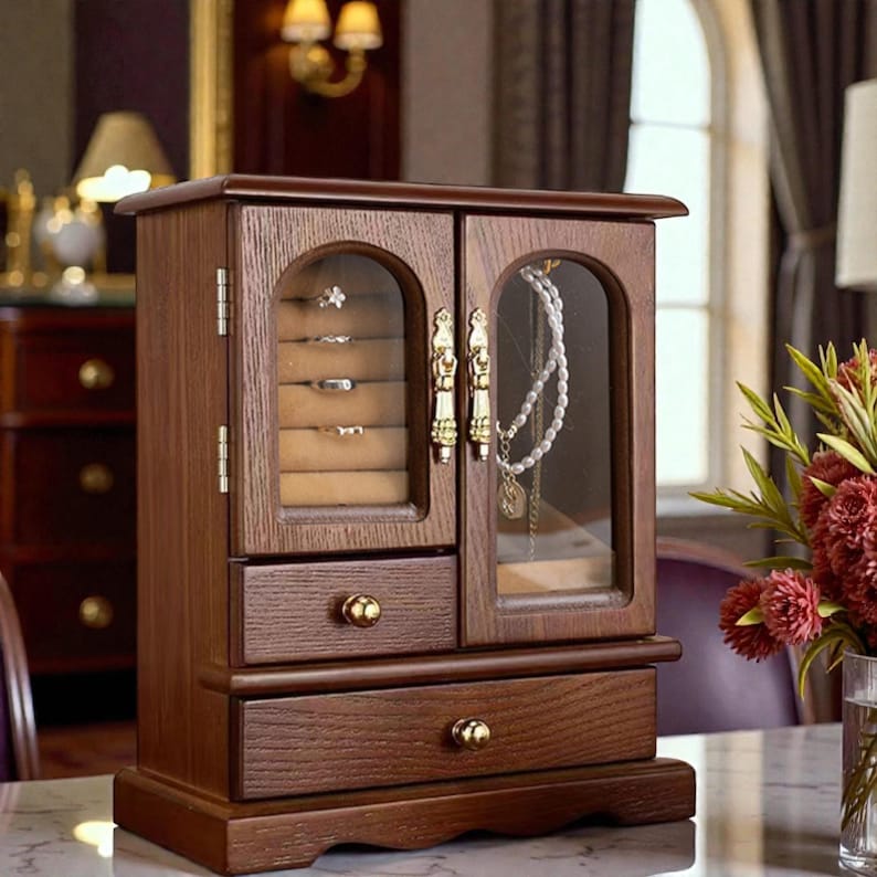 May include: A wooden jewelry box with a dark brown finish. The box has two doors with glass panels, revealing ring storage and necklace display. Two drawers are located below the doors. Gold-colored hardware accents the doors and drawers.
