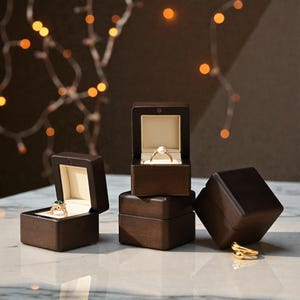 May include: Several dark brown wooden ring boxes, some open to reveal rings with gemstones and pearls. The boxes have a light-colored interior and are arranged on a light-colored surface. A string of lights is in the background.