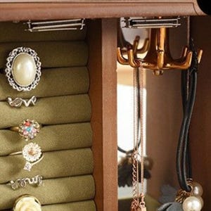 May include: A wooden jewelry box with a green velvet ring holder and a gold-toned hook for necklaces. Rings with various designs, including a pearl and floral accents, are displayed. Necklaces and a black corded necklace with pearls hang from the hooks.