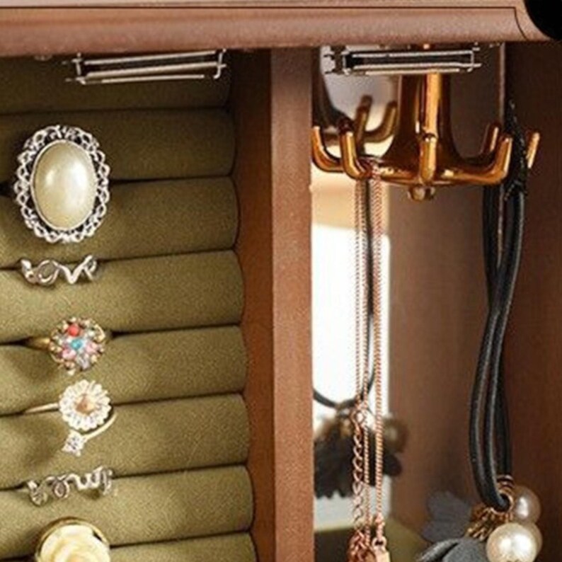 May include: A wooden jewelry box with a green velvet ring holder and a gold-toned hook for necklaces. Rings with various designs, including a pearl and floral accents, are displayed. Necklaces and a black corded necklace with pearls hang from the hooks.