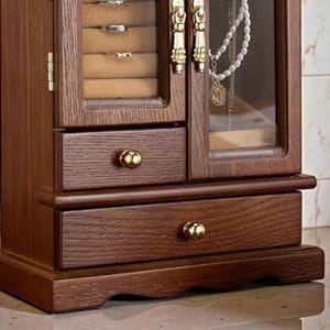 May include: A wooden jewelry armoire with a dark brown finish. It features a glass-fronted door, drawers with gold-colored knobs, and compartments for rings and necklaces. The armoire has a classic, elegant design.