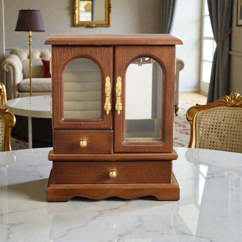 May include: A wooden jewelry box with a dark brown finish. The box has two doors with glass panels and gold-colored handles. It features a drawer and a base with a curved design. The jewelry box is sitting on a white marble surface.
