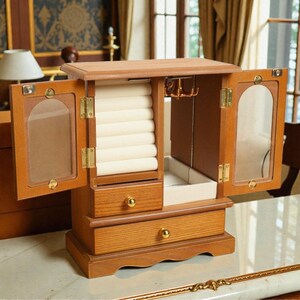 May include: Wooden jewelry box with two doors, drawers, and a lift-up top. The box is open, revealing a cream-colored interior with ring holders and hooks. The doors have glass panels. The box is made of light brown wood.