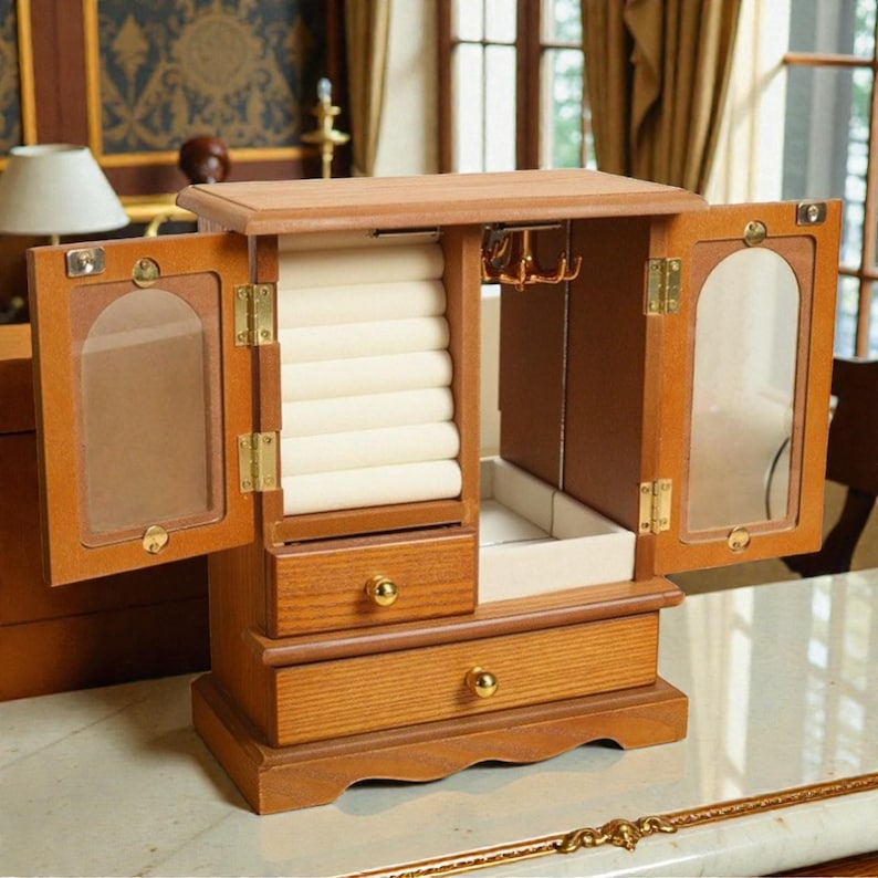 May include: Wooden jewelry box with two doors, drawers, and a lift-up top. The box is open, revealing a cream-colored interior with ring holders and hooks. The doors have glass panels. The box is made of light brown wood.
