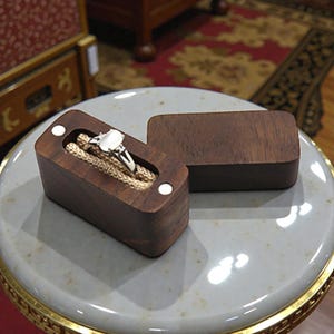 May include: A wooden ring box with a silver ring with a white gemstone inside. The box is rectangular, made of dark brown wood, and has a small, rectangular lid. The ring is set on a beige cushion inside the box.