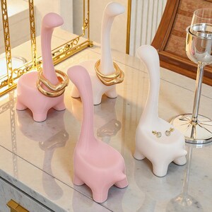 May include: Four dinosaur-shaped ring holders in pink and white. Two holders display gold rings, while another holds earrings. The holders are on a reflective surface, with a gold-colored decorative element in the background.