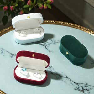 May include: Three velvet ring boxes in blue, burgundy, and green, each with a gold clasp. Two boxes are open, displaying rings with clear and blue gemstones. The boxes sit on a marble-patterned surface.