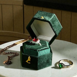 May include: A dark green velvet hexagonal ring box with a gold clasp, open to reveal a red gemstone ring. A gold necklace and a gold ring with a green gemstone are also visible on a white surface. The box is a luxurious jewelry display.