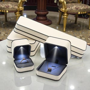 May include: Cream-colored jewelry boxes with black trim, some open to reveal a ring and a necklace. The boxes have built-in lights that illuminate the jewelry. The boxes are rectangular and square-shaped.