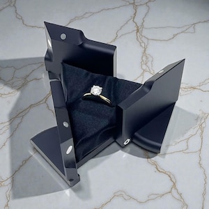 Peut inclure: Un écrin à bagues noir et géométrique contenant une bague en or ornée d'une grosse pierre précieuse transparente. La bague repose sur un tissu noir à l'intérieur de la boîte ouverte. La boîte a un design moderne et angulaire.