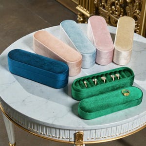 Puede incluir: Una colección de joyeros de terciopelo en varios colores, incluyendo turquesa, rosa y dorado, dispuestos sobre una mesa con tapa de mármol. Varias cajas están abiertas, mostrando anillos con piedras preciosas brillantes. Las cajas están diseñadas para anillos.