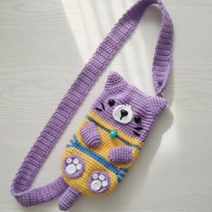 Op de afbeelding: Handgemaakte gehaakte crossbody tas in de vorm van een kat, in paars, geel en blauw. De tas heeft een lange riem, een kattengezicht met zwarte ogen en snorharen en pootafdrukken op de voorkant. De tas is ongeveer 25 cm lang.