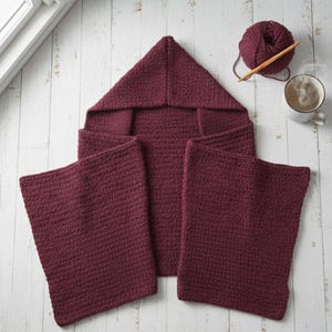May include: A burgundy hooded baby blanket with matching bibs, all hand-knitted. A ball of burgundy yarn and a knitting needle are next to a mug of coffee. The blanket is folded and displayed on a white wooden surface.