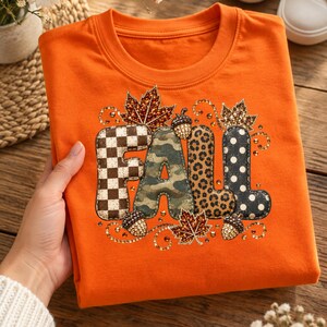May include: A bright orange t-shirt featuring the word FALL in large letters. Each letter has a different pattern: brown and white checkerboard, green camouflage, leopard print, and black with white polka dots, accented by rhinestones and leaves.