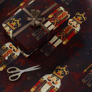 May include: A dark red and black wrapping paper featuring a repeating nutcracker pattern with gold accents. A rectangular gift box is wrapped in the same paper, tied with a brown ribbon bow, accompanied by a roll of paper, scissors, and tape.