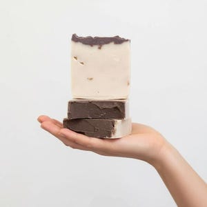 Può includere: Tre saponette fatte a mano impilate. La saponetta superiore è beige chiaro con la parte superiore marrone scuro, la saponetta centrale è marrone scuro e la saponetta inferiore è beige chiaro con la parte inferiore marrone scuro. I saponi sono tenuti in una mano su sfondo bianco.