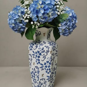 May include: A white ceramic vase with a hexagonal shape, decorated with blue floral patterns and small gold accents, filled with a bouquet of blue hydrangea flowers, white baby's breath, and green eucalyptus leaves.