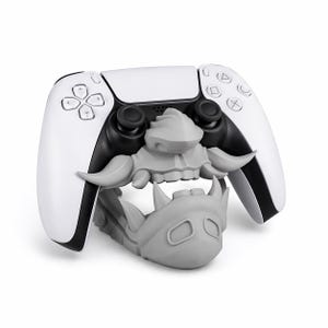 Puede incluir: Un mando de PlayStation 5 blanco y negro con un soporte gris. El soporte está diseñado con una cara estilizada de monstruo gris. El mando tiene joysticks y botones negros.
