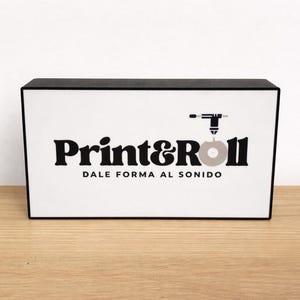 Könnte beinhalten: Rechteckige Box mit schwarzem Deckel und Rahmen, mit dem Text "Print&Roll" in fetter, schwarzer Schrift. Darunter der Satz "DALE FORMA AL SONIDO" und eine Grafik einer Bohrmaschine und einer Schallplatte.