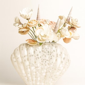 Könnte beinhalten: Eine dekorative, muschelförmige Vase, gefüllt mit einem Blumenarrangement. Das Arrangement umfasst weiße, beige und pfirsichfarbene Muscheln und blütenförmige Muscheln. Die Vase hat eine gesprenkelte, perlmuttartige Oberfläche.