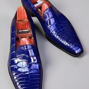 Op de afbeelding: Een paar blauwe leren herenschoenen met een krokodillenleerpatroon. De schoenen hebben een spitse neus en een houten schoenspanner aan de binnenkant. Op een grijze achtergrond.